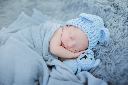 Portrait of twenty seven day old newborn asian baby boy in knitted blue hat with teddy bear sleeping...