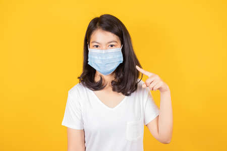Portrait of Asian teenager  wearing and pointing medical face mask to protect virus wears white t-shirt on yellow backgroundの写真素材