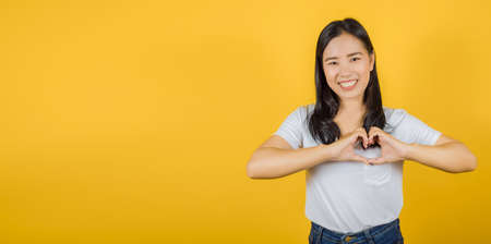 Portrait of young beautiful Asian woman in love making heart with fingers on yellow background, copy spaceの写真素材