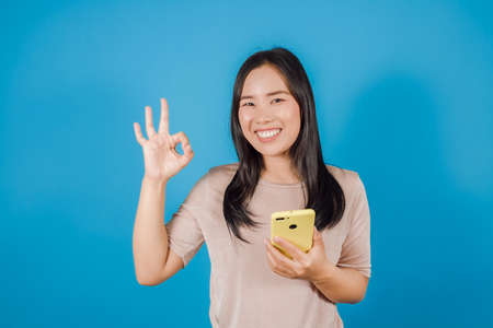 Portrait of happy Asian woman wears nude t-shirt showing ok-sign holding mobile phone or smartphone isolated on blue backgroundの写真素材