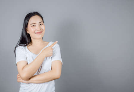Portrait of smiling Asian woman with long dark hair wears white t-shirt pointing finger to the side isolated on gray background with copy spaceの写真素材