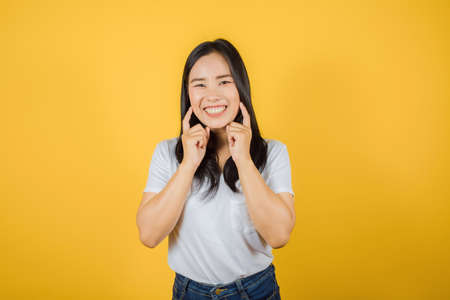 Portrait of smiling Asian woman with long dark hair wears white t-shirt keeps index fingers on cheeks isolated on yellow backgroundの写真素材