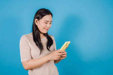 Portrait of smiling Asian woman wears nude t-shirt using a smartphone isolated on blue backgroundの写真素材