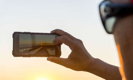 Man with glasses taking photo with mobile phone, sunset light.の写真素材