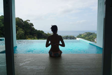 Slim young woman in bikini sitting on the edge of the swimming pool and looking at sea view.の写真素材