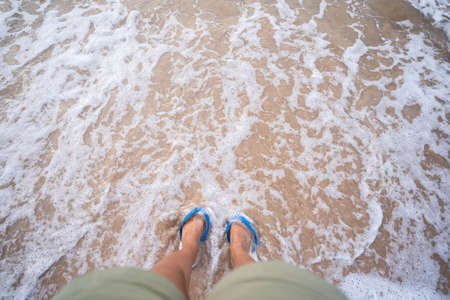 A woman wearing white sandals Stand on the beach with white waves.の写真素材