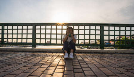 Asian woman wearing a surgical mask sits on a bridge, hands holding her head due to unemployment stress, behind a river and sunset.の写真素材