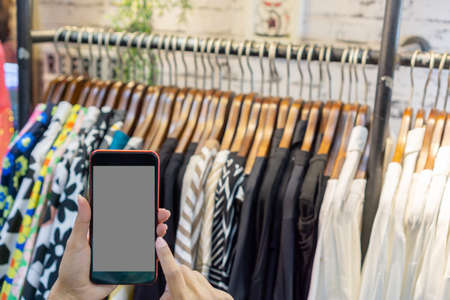 Woman's hand holding a phone in clothing store, blurred clothing store background, shopping online concept.の写真素材
