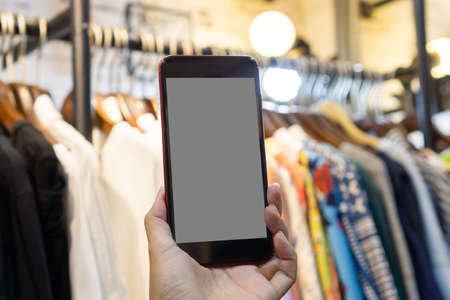 Woman's hand holding a phone in clothing store, blurred clothing store background, shopping online concept.の写真素材