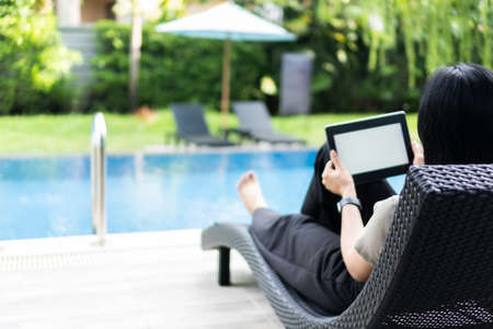 Female tourist sits on a chair by the pool at the hotel, holding a tablet with isolated screen.
