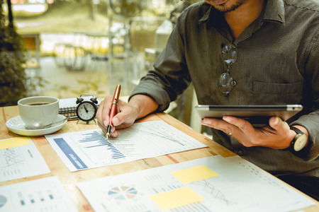Hand right of businessman holding a pen and writing graphs paper. There is cup of coffee and clock...