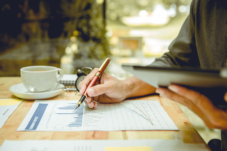 Hand right of businessman holding a pen and writing graphs paper. There is cup of coffee and clock on wooden table. His left hand holding a digital tablet.の写真素材