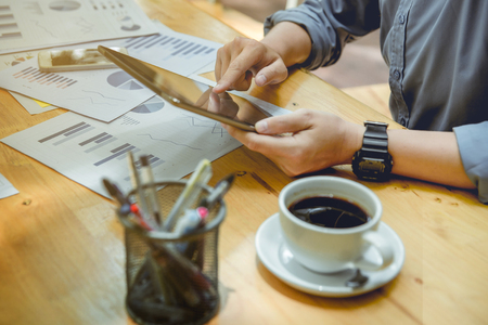 hands of officer holding and touching digital tablet. There is many charts papers, cup of coffee and smart phone on wooden table.の写真素材
