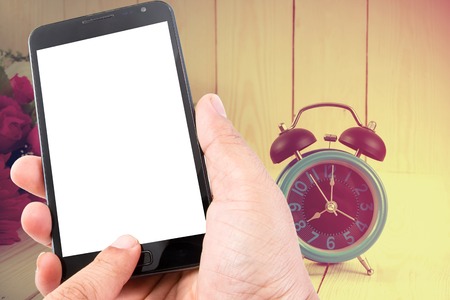 Man hand hold blank touch screen smart phone on vintage wood background and flowers with retro alarm clock.の写真素材