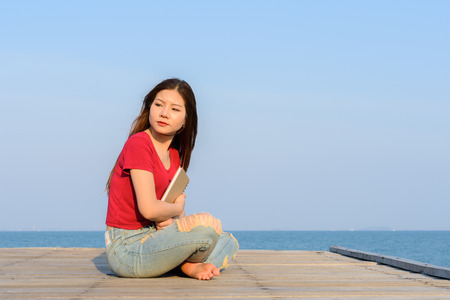 woman sitting holding a books on the wooden bridge sea and blue sky in backgroundの写真素材