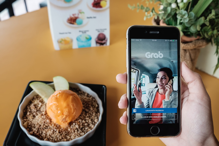 CHIANG MAI,THAILAND - JAN 7,2017 : A MAN hand holding Grab app showing on iphone 6s  with computer laptop and ear podon wood desk in coffee shop. Grab is smartphone app-based transportation networkのeditorial素材