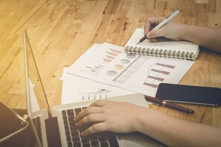 Closeup business man hand holding pen point notebook with using laptop computer and smartphone and stock maket chart on wood desk in office. vintage effect.の写真素材