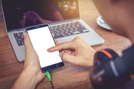 A man hand holding smart phone and use laptop computer on wood desk. Smartphone with blank screen and can be add your texts or others on smart phone. vintage effect.の写真素材