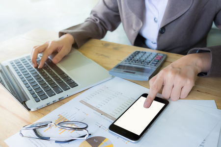 Business womanhand pointing finger on smartphone and using laptop computer with calculator and stock market chart on wood desk.の写真素材