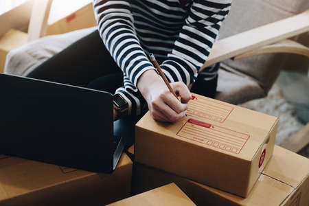 Online selling business idea, beautiful girl writing customer address in front of parcel delivery box and using computer to enter tracking and trace number of parcels for customers.の写真素材