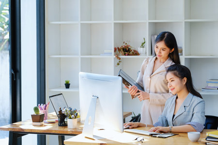 Consultation, discussion, marketing and investment concept, female employee holding folder and colleague using a computer monitor to draw conclusions and assess investment risks for the company.の写真素材
