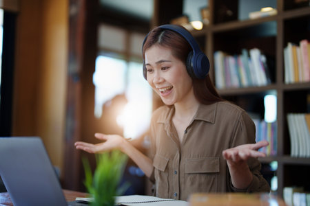 Portrait of a smiling Asian teenage girl wearing headphones and using a computer for online video conferencing in a library.の写真素材