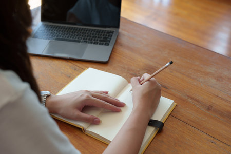 Portrait of a teenage Asian woman using a computer and notebook to study online via video conferencing on a wooden desk in library.の写真素材