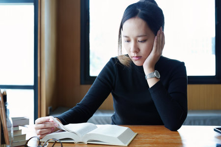 A portrait of a young Asian woman reading textbooks shows boredom on a wooden desk in library..の写真素材