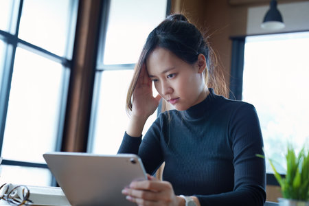 Portrait of a teenage Asian woman using a tablet computer and books to study online via video conferencing on a wooden table in the library.の写真素材