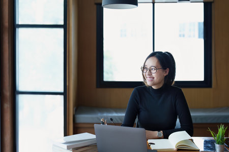 Portrait of a teenage Asian woman wearing glasses using computer laptop, headphones and using a laptop to study online via video conferencing on a wooden library table.の写真素材