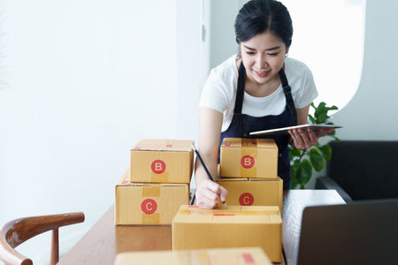 Starting small business entrepreneur of independent Asian woman smiling using tablet computer with cheerful success of online marketing package box items and SME delivery concept.の写真素材