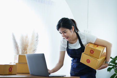 Starting small business entrepreneur of independent Asian woman smiling using computer laptop with cheerful success of online marketing package box items and SME delivery concept.の写真素材
