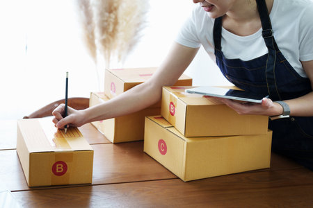 Starting small business entrepreneur of independent Asian woman smiling using tablet computer with cheerful success of online marketing package box items and SME delivery concept.の写真素材