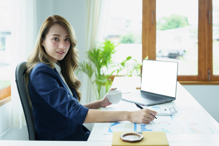 Portrait of an Asian business woman drinking coffee while working with a computer and financial statements documents on her desk.の写真素材