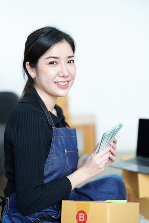 Starting small business entrepreneur of independent Asian woman smiling and holding money using laptop computer with cheerful success of online marketing package box items and SME delivery concept.の写真素材