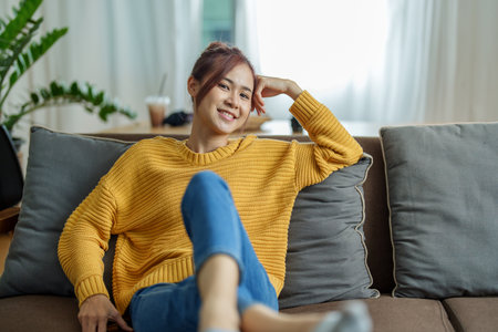 A portrait of a young Asian woman resting with a smiling face on the sofa by the window of the house.の写真素材