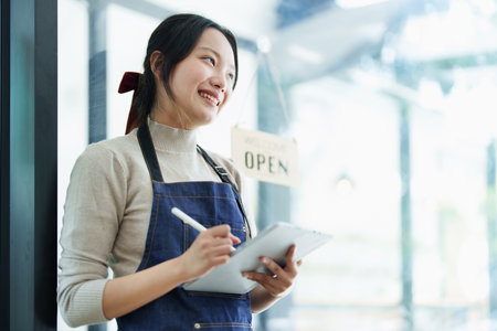 Starting and opening a small business, a young Asian woman showing a smiling face holding a tablet in an apron standing in front of a coffee shop bar counter. Business Owner, Restaurant, Cafe concept.の写真素材