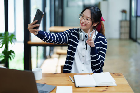Portrait of a teenage Asian woman using a smartphone, wearing headphones and using a notebook to study online via video conferencing on a wooden desk in library.の写真素材