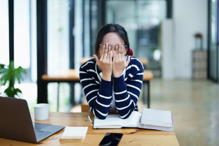 A portrait of a young Asian woman using a computer, wearing headphones and using a notebook to study online shows boredom and pain from video conferencing on a wooden desk in library.の写真素材