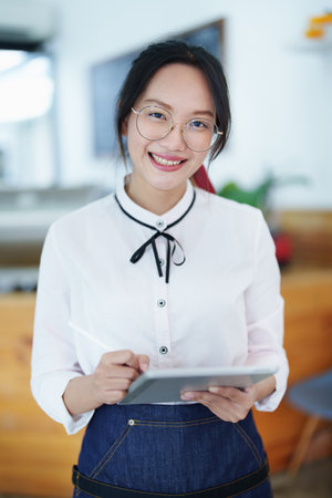 Starting and opening a small business, a young Asian woman showing a smiling face holding a tablet in an apron standing in front of a coffee shop bar counter. Business Owner, Restaurant, Cafe concept.の写真素材