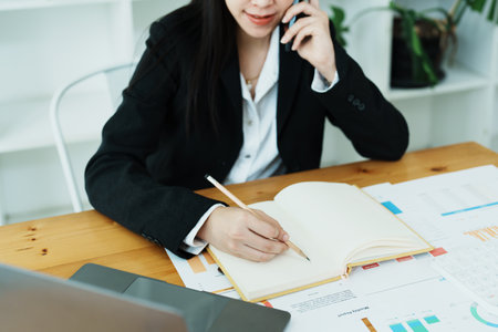 Portrait of a young Asian woman showing a smiling face as she uses her phone, computer and financial documents on her desk in the early morning hours.の写真素材