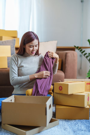 Portrait of a small startup Asian female entrepreneur SME owner picking up shirt before packing it...