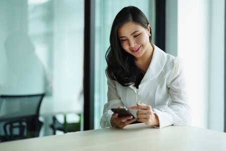Portrait of a young Asian woman sitting at a desk using her phone.の写真素材