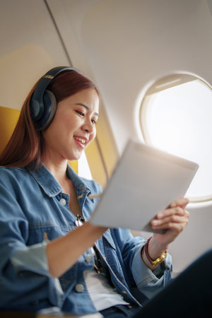 Attractive portrait of an Asian woman sitting in a window seat in economy class using tablet and...