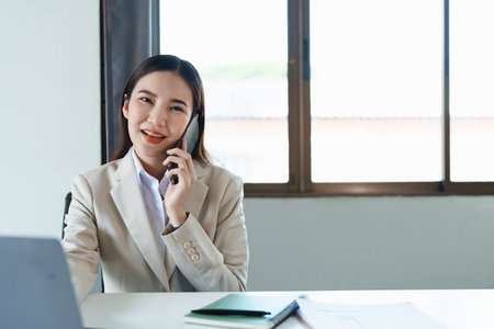 Portrait of a young Asian woman showing a smiling face as she uses her phone, computer and financial documents on her desk in the early morning hours.の写真素材