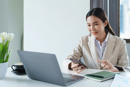 Portrait of a young Asian woman showing a smiling face as she uses her phone, computer and financial documents on her desk in the early morning hours.の写真素材