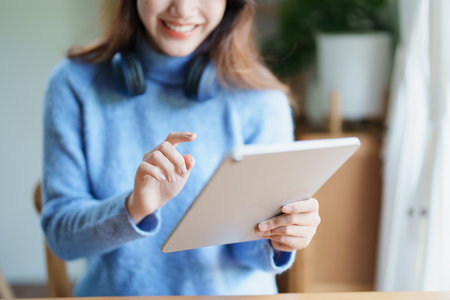 Portrait of a teenage Asian woman using a tablet, wearing headphones to study online via video...