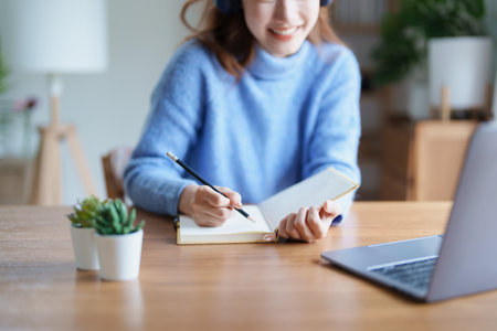Portrait of a teenage Asian woman using a computer, wearing headphones and using a notebook to study...