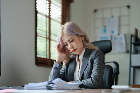 Portrait of a young Asian woman showing a serious face as she using financial documents and computer laptop on her desk in the early morning hours.の写真素材