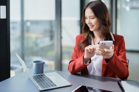 Portrait of a young Asian woman showing a smiling face as she uses her phone, computer and financial documents on her desk in the early morning hours.の写真素材